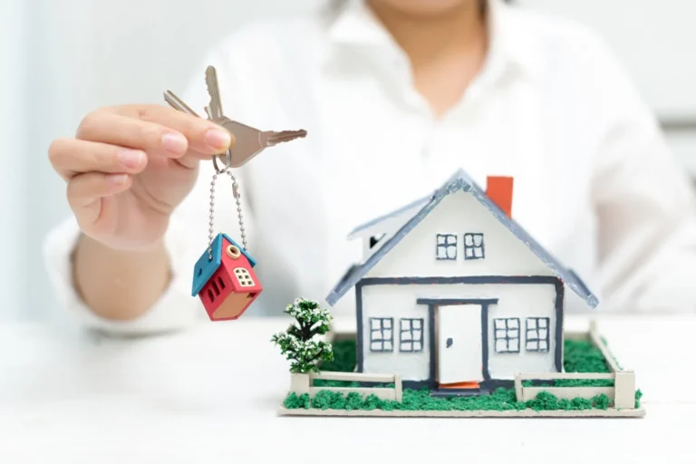 มือถือกุญแจบ้านกับบ้านจำลอง / Holding house keys next to a model house